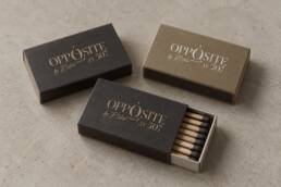Charcoal and muted gold matchbox mockups with Opposite by Botrini at 507 branding